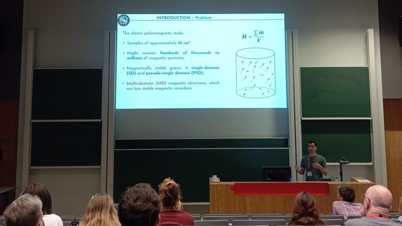 Presenter (brown-skinned male in a green t-shirt) behind lectern next to presentation screen with introduction slide. Slide contains classic paleomagnetic studies that use samples of approximated 10 cm³ with hundreds of thousands to millions of individual magnetic particles. Also seen are the backs of the heads of 8 audience members.