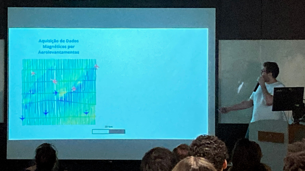 Presenter (light-skinned male in a white t-shirt) next to presentation screen with a slide containing a diagram about aeromagnetic surveys. Also seen are the backs of the heads of 7 audience members.