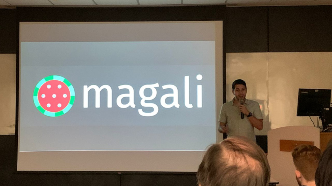 Presenter (light-skinned male in a green shirt) next to presentation screen with a slide containing a Magali`s logo (a watermelon-like figure in which its seeds resemble magnetic dipoles, followed by the project's name). Also seen are the backs of the heads of 3 audience members.