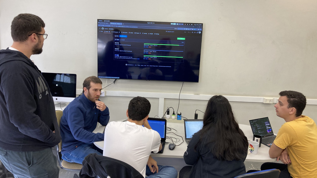 Four people sitting around a table with laptops open and one person standing. Behind them is a TV on a wall with a browser open to the website GitHub.